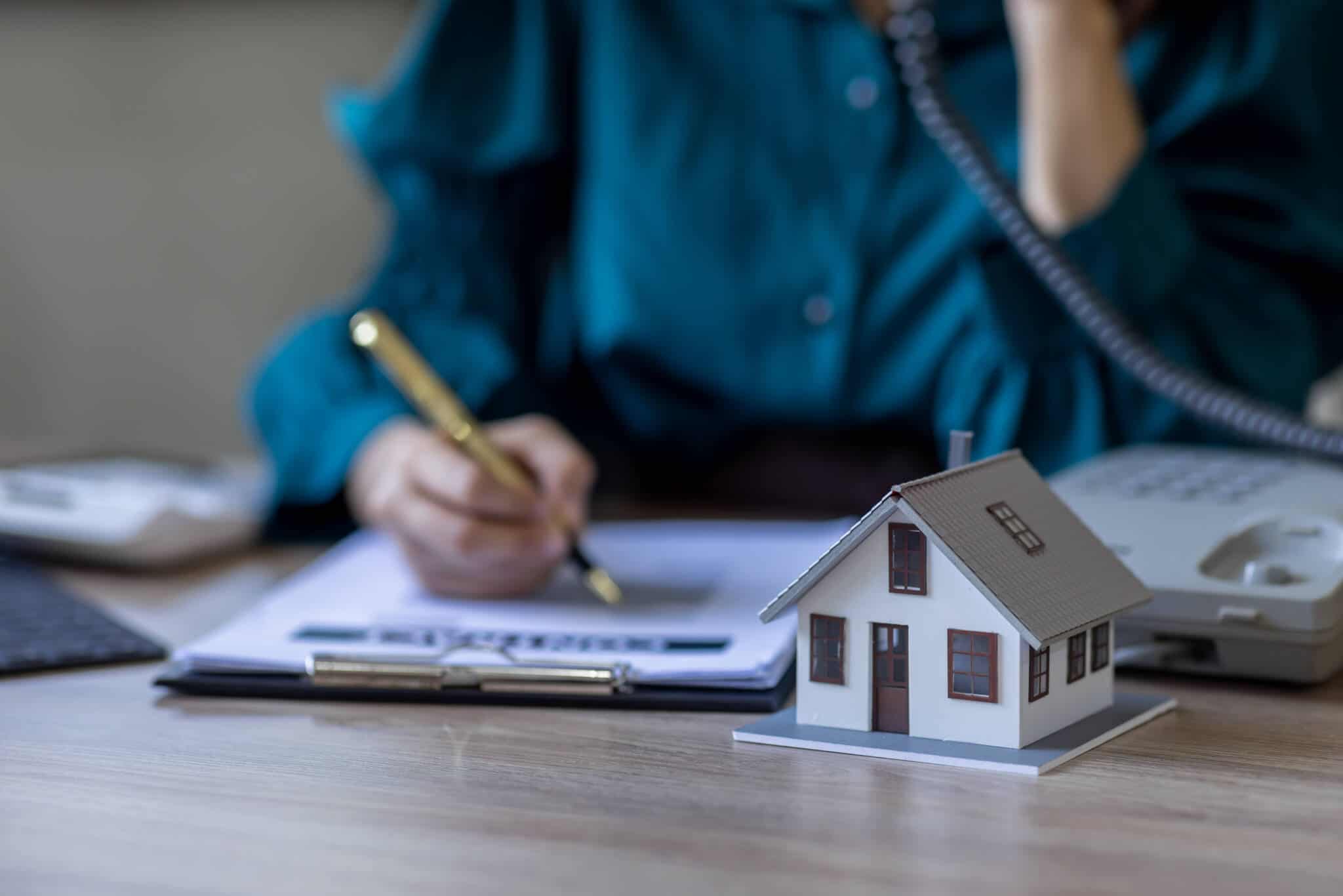 Woman sitting at the desk signing a document with a wooden house next to it, symbolizing the change to a fixed interest rate on mortgage loans