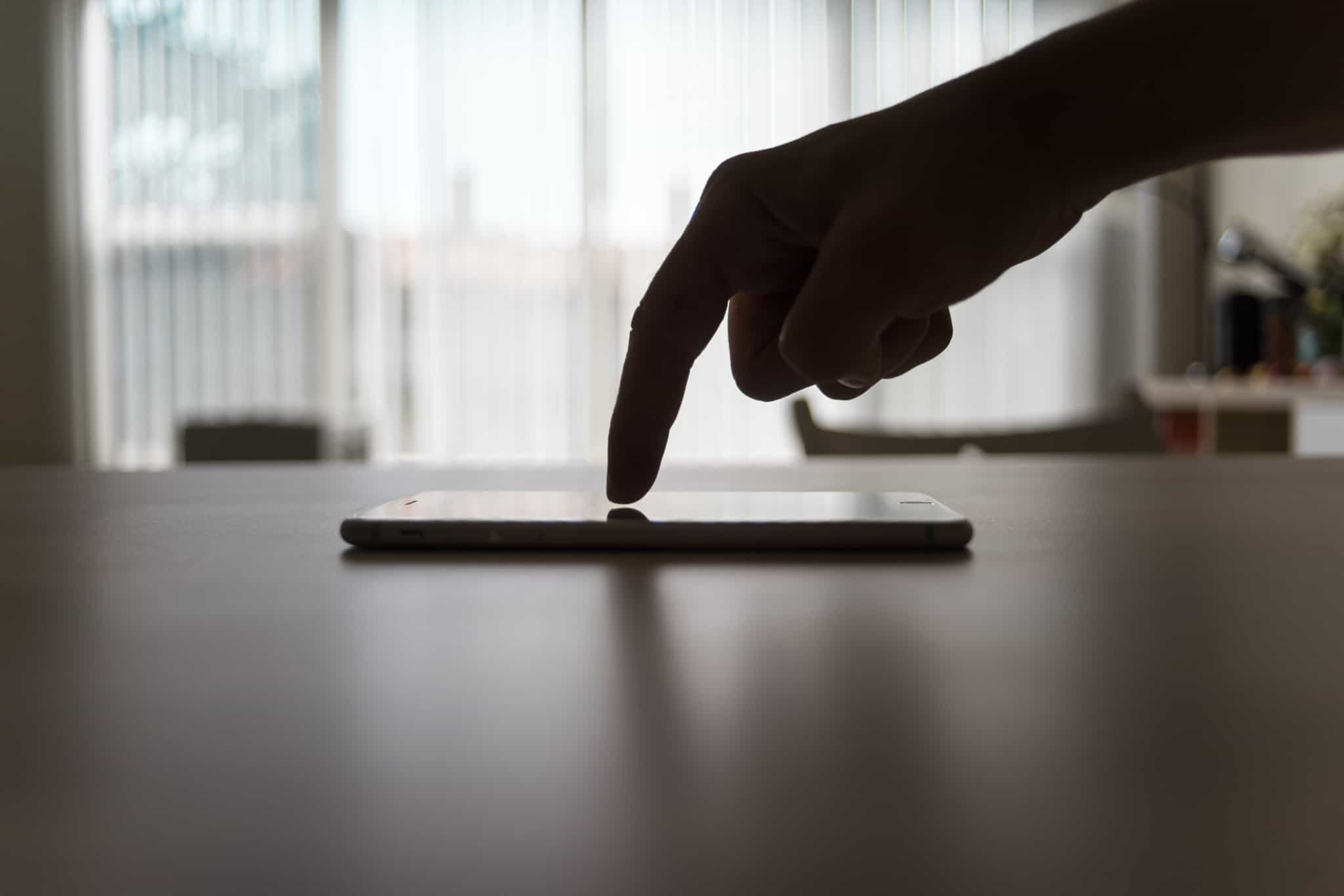 in a backlit image, a finger runs across the screen of a mobile phone, on top of a table, while accessing the bank account on the mobile phone